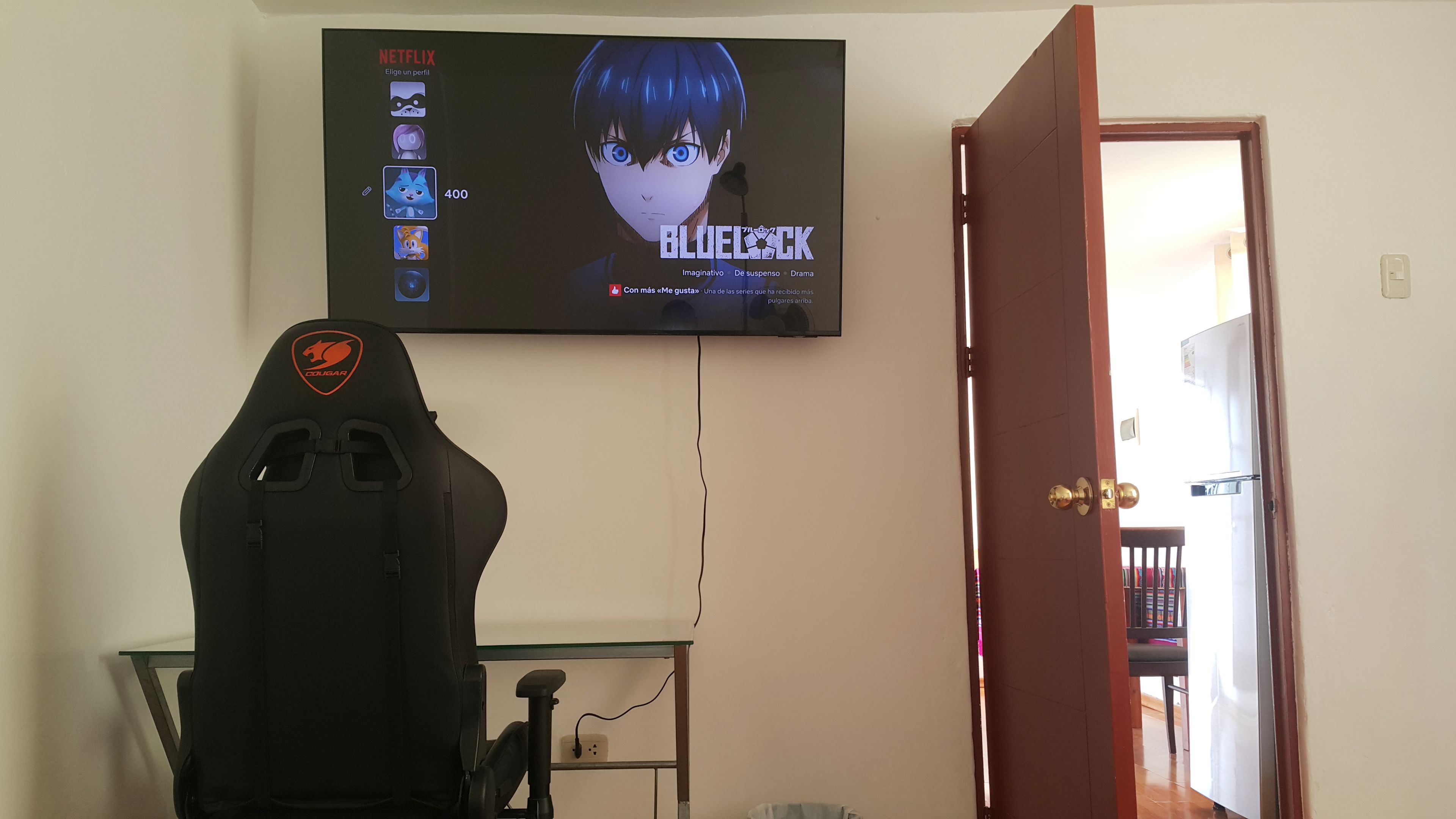 Una habitación con una televisión en la pared mostrando un programa, un sillón gamer y una puerta que da a otra habitación.