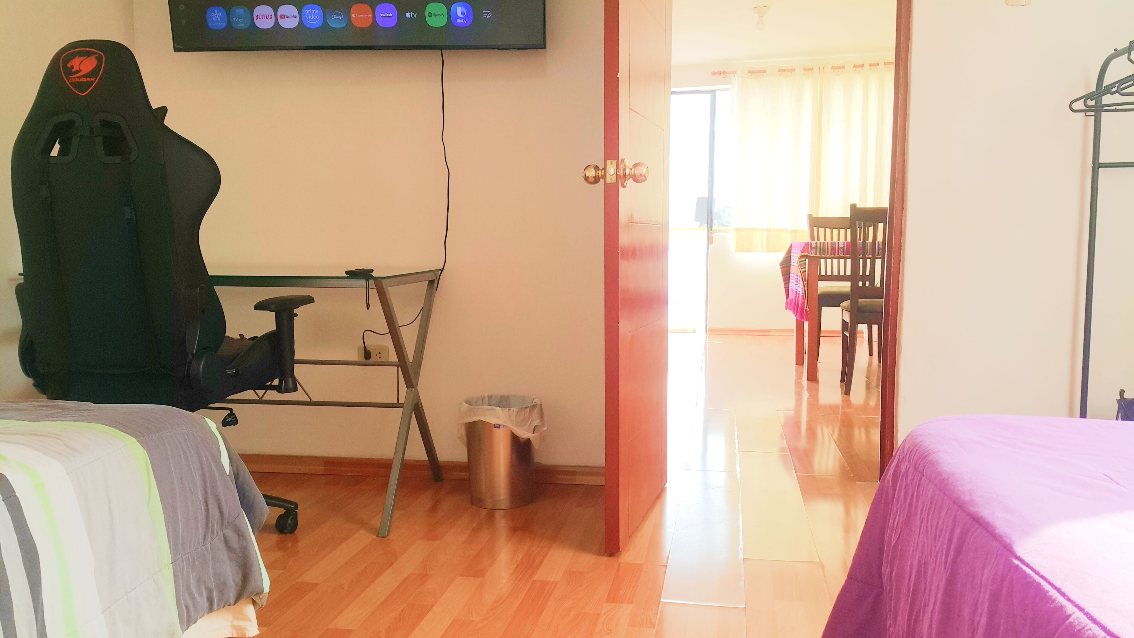 A hotel room with a bed, a desk with a chair, a trash bin, and a television mounted on the wall displaying various streaming services.