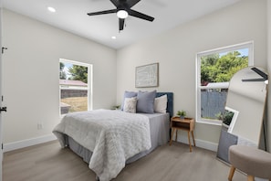 Une chambre à coucher avec un lit, un ventilateur de plafond, une table de chevet, une chaise et deux fenêtres.