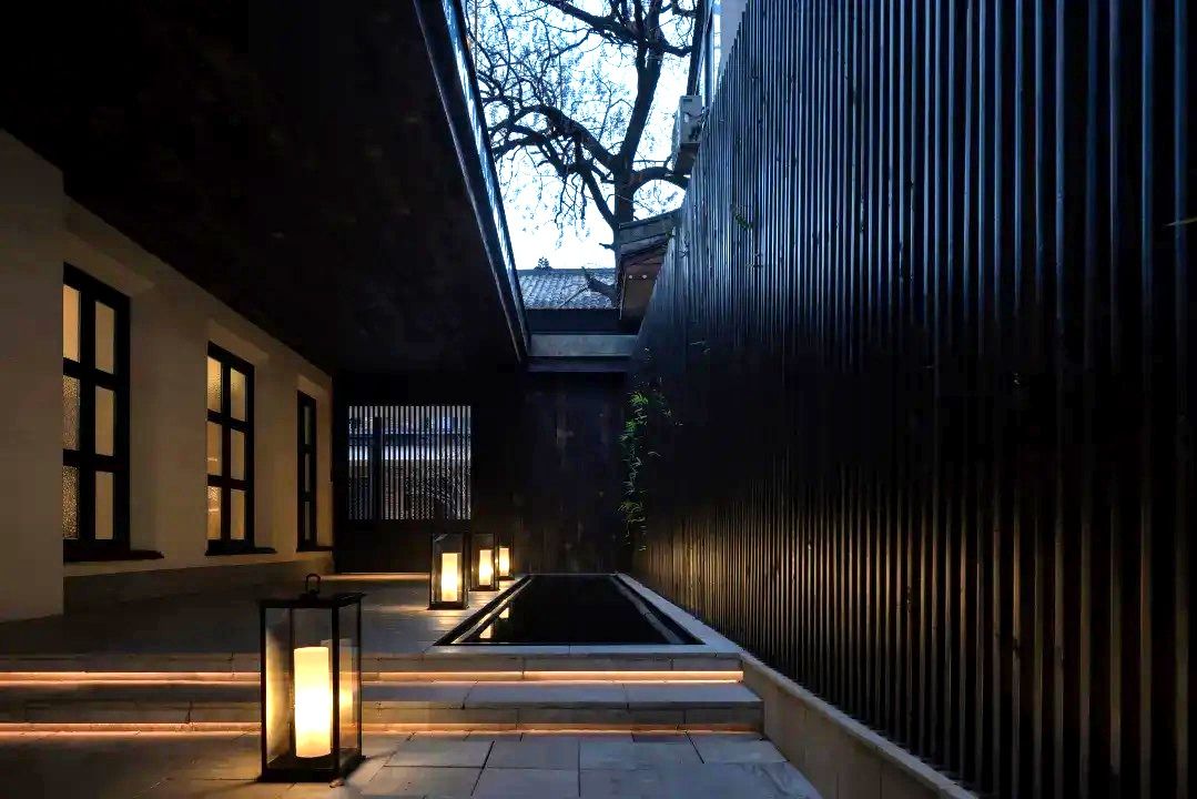 A narrow pathway with steps leading to a water feature, flanked by walls with windows and lanterns.