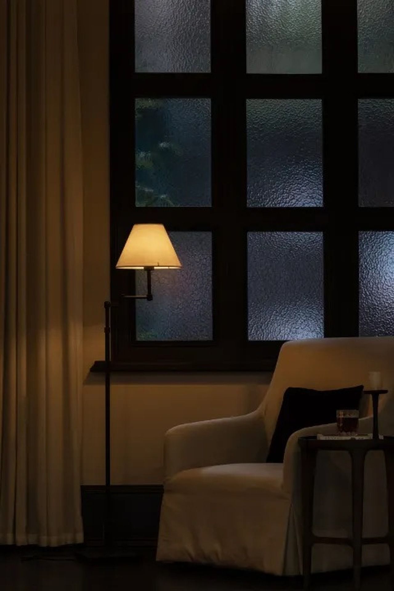 Quarto com uma cadeira, uma mesa, uma janela com vidro fosco e um abajur.