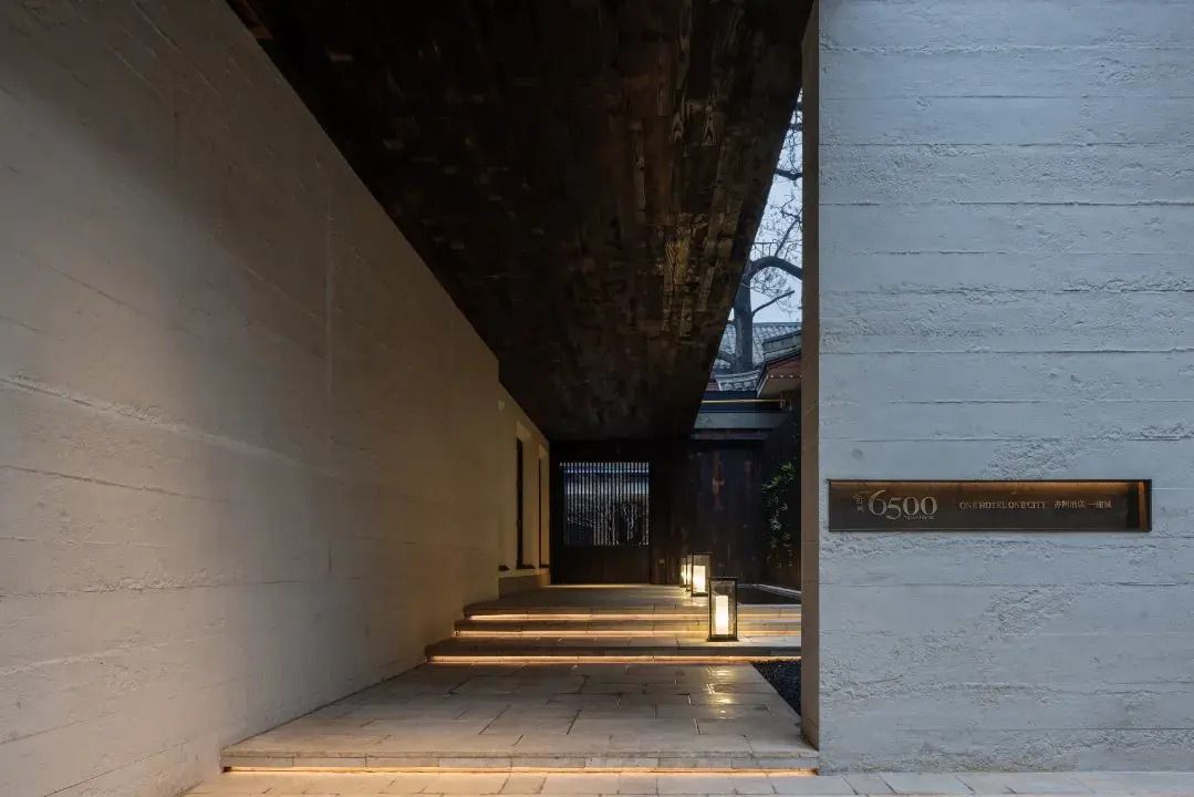 A corridor with steps leading to a door, a sign with the number 6500, and a light fixture on the wall.
