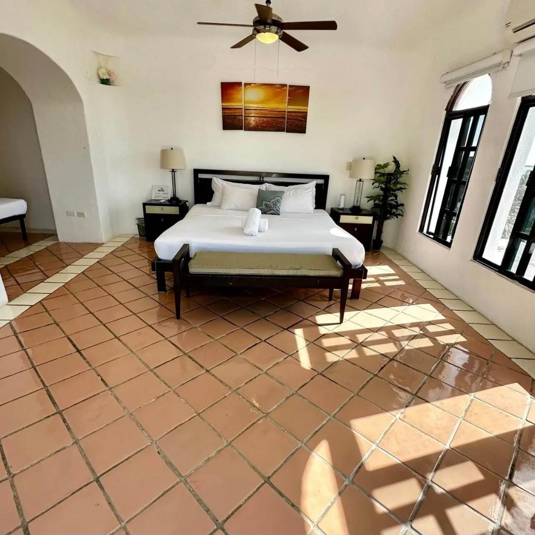 Une chambre d’hôtel avec un lit, un ventilateur de plafond, un tableau, une table de chevet, une lampe, une plante en pot et une fenêtre.