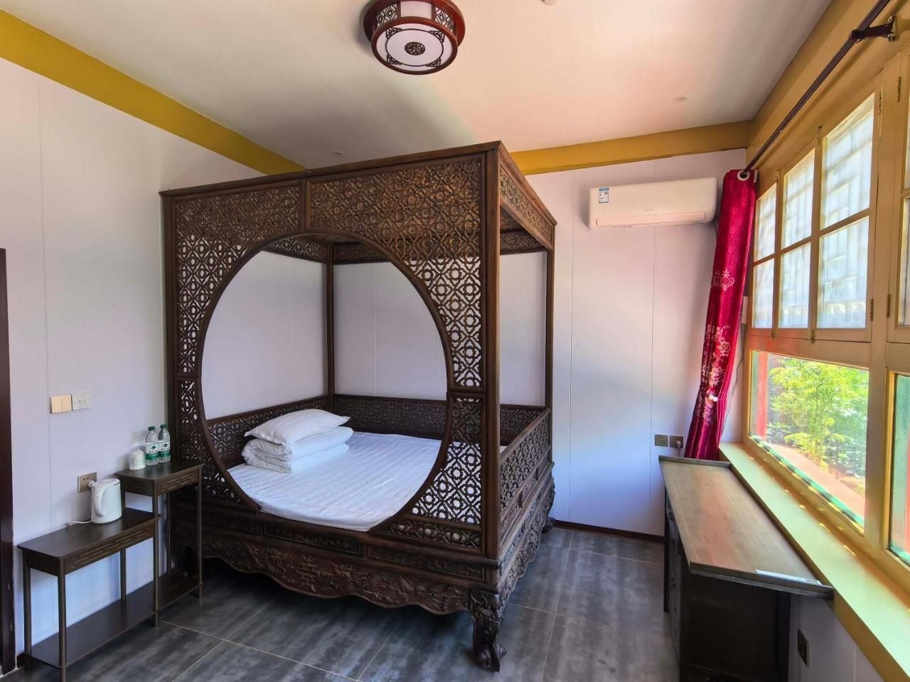 Una cama con dosel y detalles de madera tallada, una mesita de noche con hervidor, y una ventana con cortina roja.