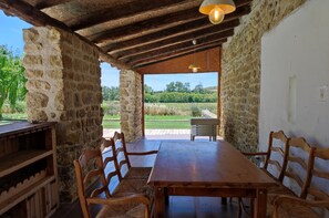 A rustic wooden dining area with a table and chairs, stone walls, and a view of a green landscape.