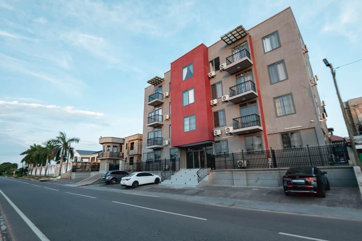 A multi-story residential building with balconies, a red accent wall, and parked cars on the street.