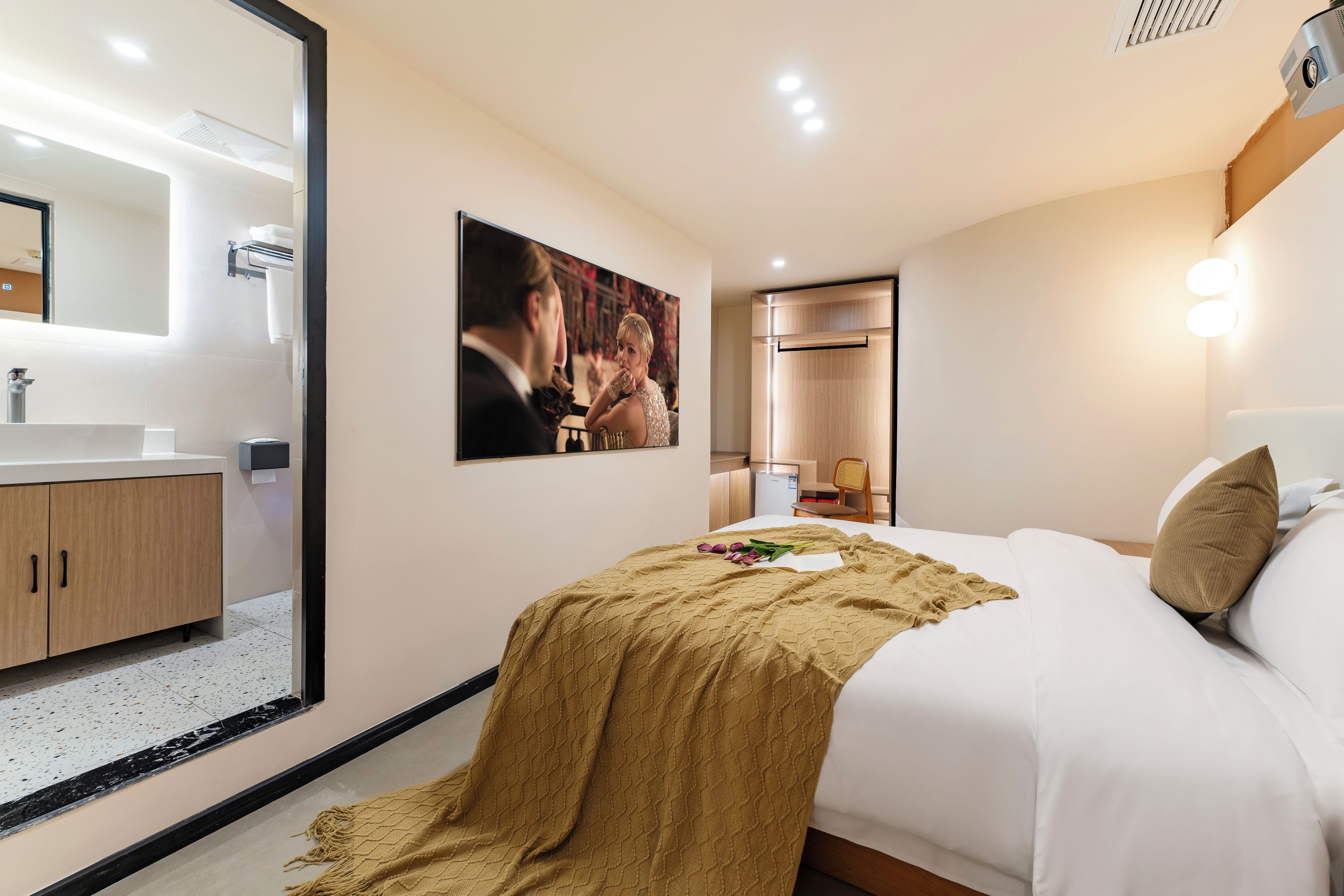 A hotel room with a large mirror, a bed with a beige blanket, a picture on the wall, and a small table with a lamp.