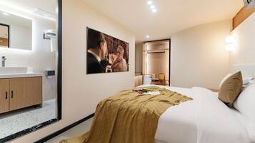 A hotel room with a large mirror, a bed with a beige blanket, a picture on the wall, and a small table with a lamp.