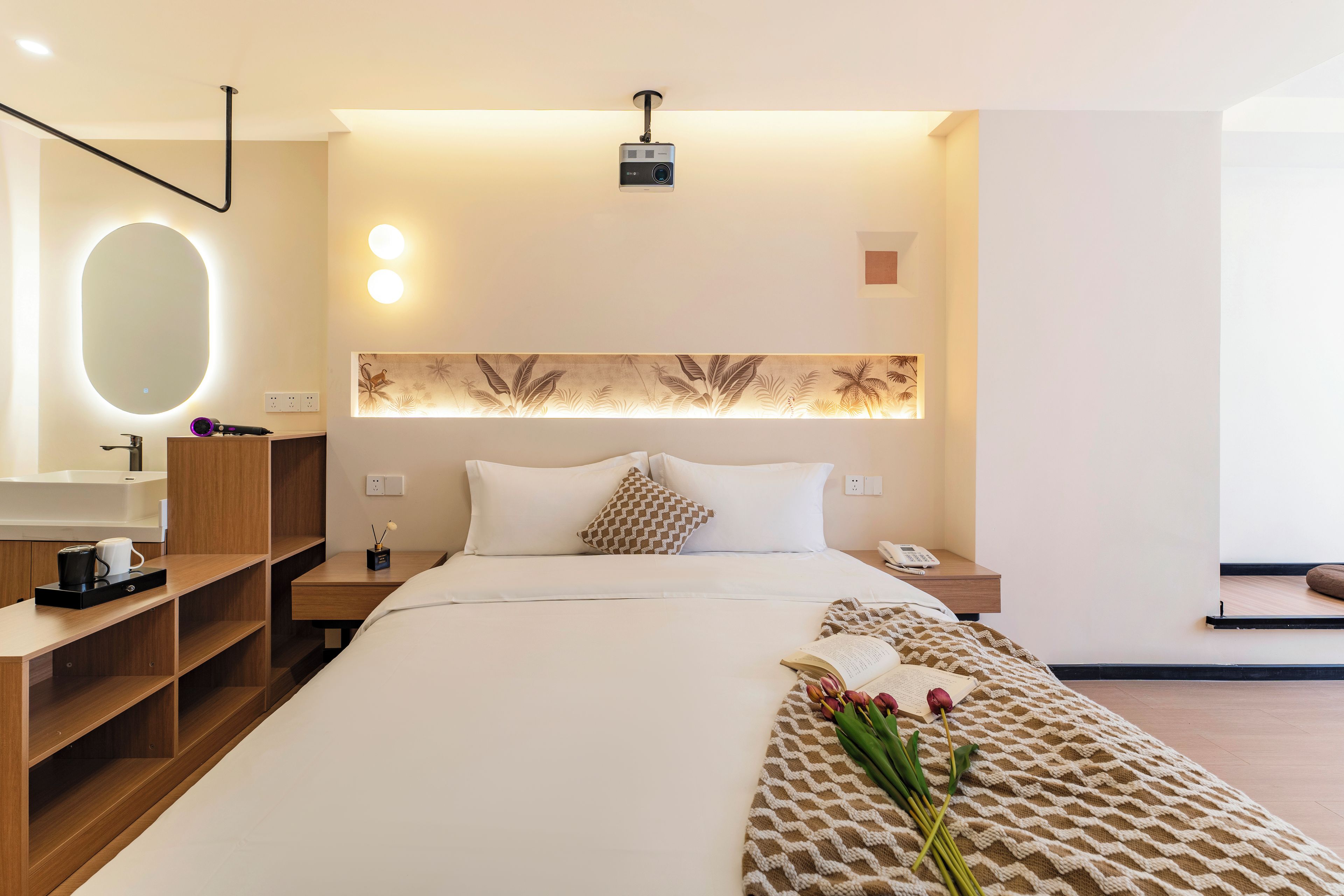 Una camera d'albergo con un letto, comodini, una scrivania, uno specchio, un proiettore a parete e un pannello decorativo.