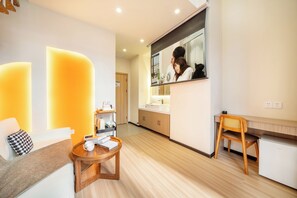 A modern hotel room with a sofa, a wooden side table, a small round table, a mirror, a kitchenette, and a wooden chair.