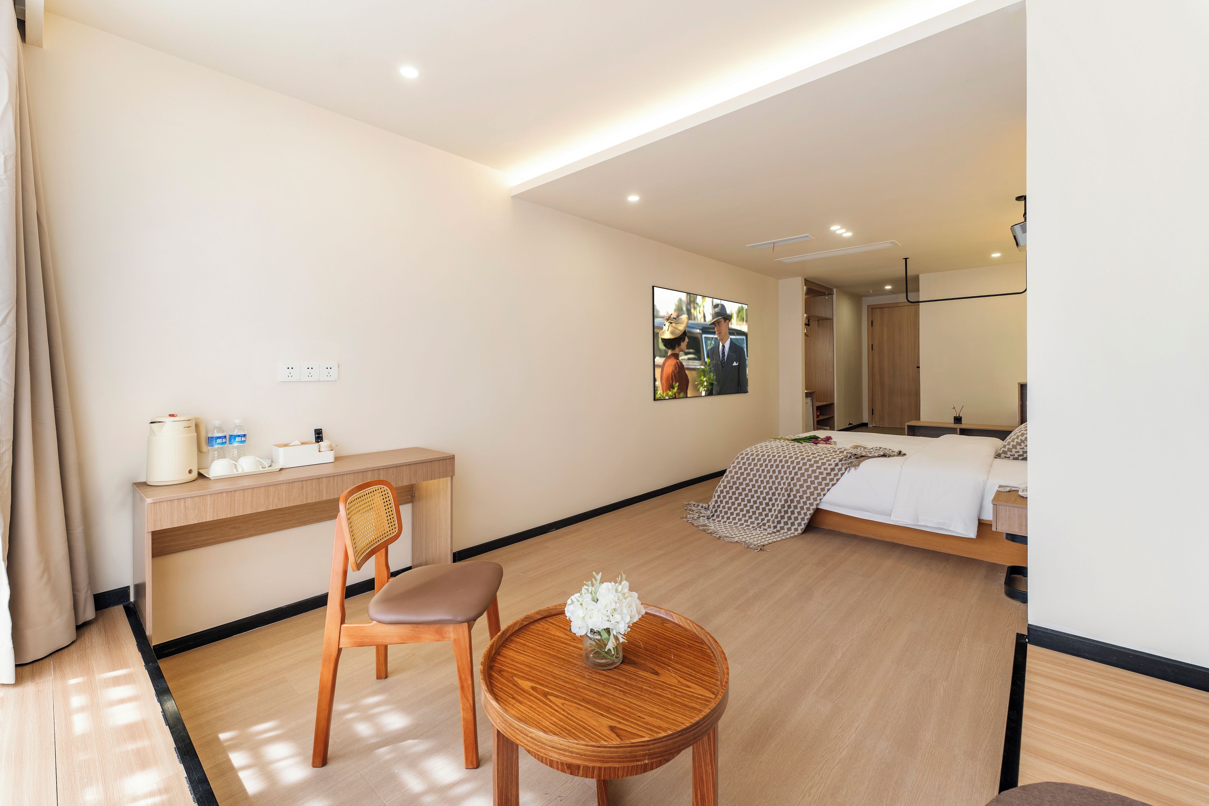 A hotel room with a bed, a desk, a chair, a small table with a vase of flowers, and a wall-mounted television.