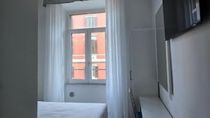 Hotellihuone, jossa on sänky, ikkuna verhoilla, seinälle asennettu televisio ja näkymä ulkona olevaan rakennukseen.