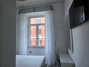 Une chambre d’hôtel avec un lit, une fenêtre avec des rideaux, un téléviseur fixé au mur et une vue sur un bâtiment à l’extérieur.
