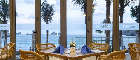 A dining area with wicker chairs and a table set for two, overlooking a body of water with palm trees and a view of the sea.