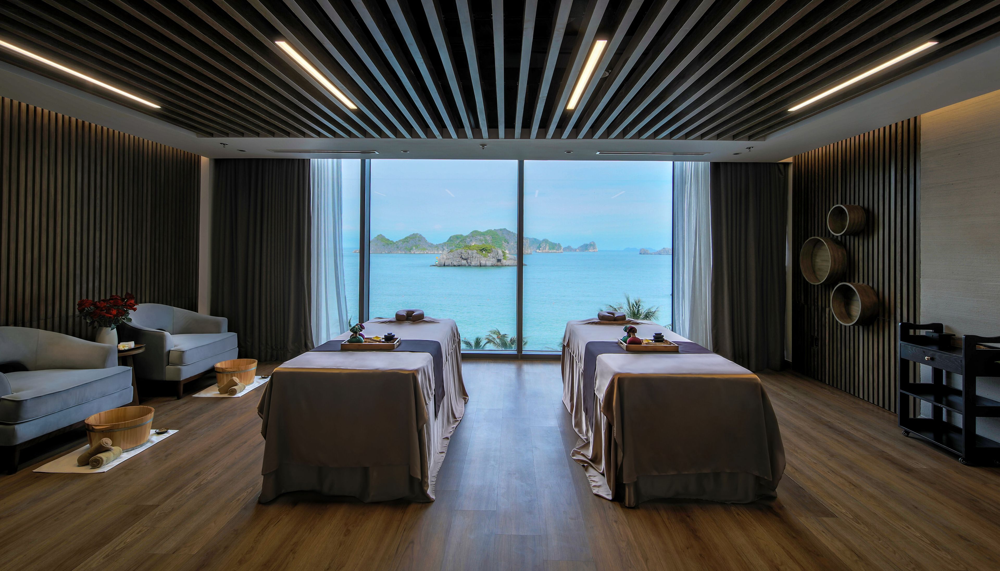 A spa room with a large window, two massage tables, a sofa, and wooden flooring.