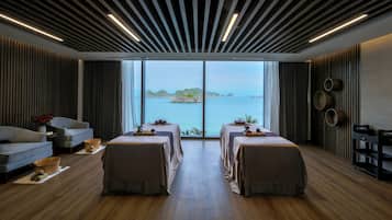 A spa room with a large window, two massage tables, a sofa, and wooden flooring.