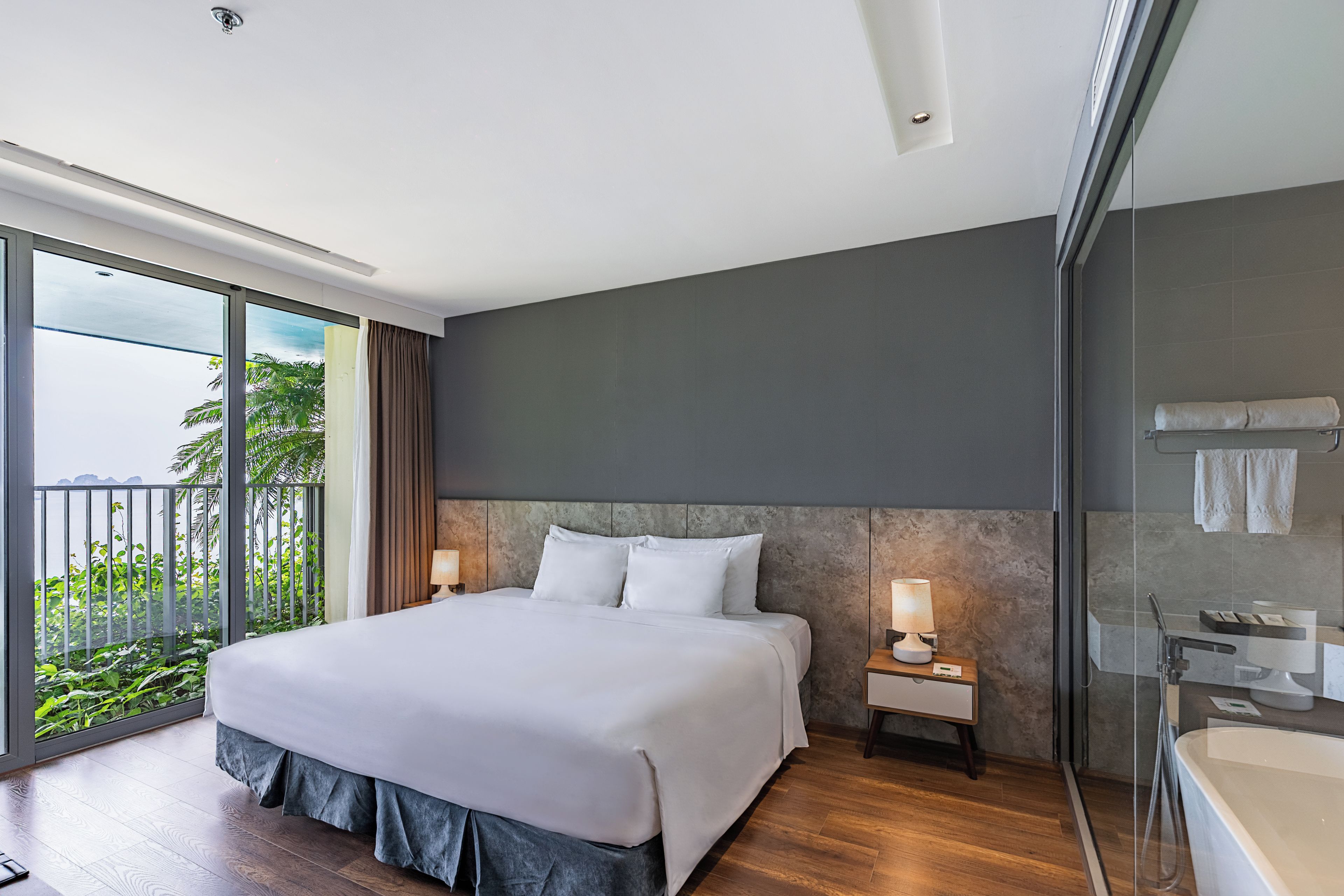 Ein modernes Hotelzimmer mit einem großen Bett, einer Glasdusche und einem Balkon mit Blick auf Grünflächen.