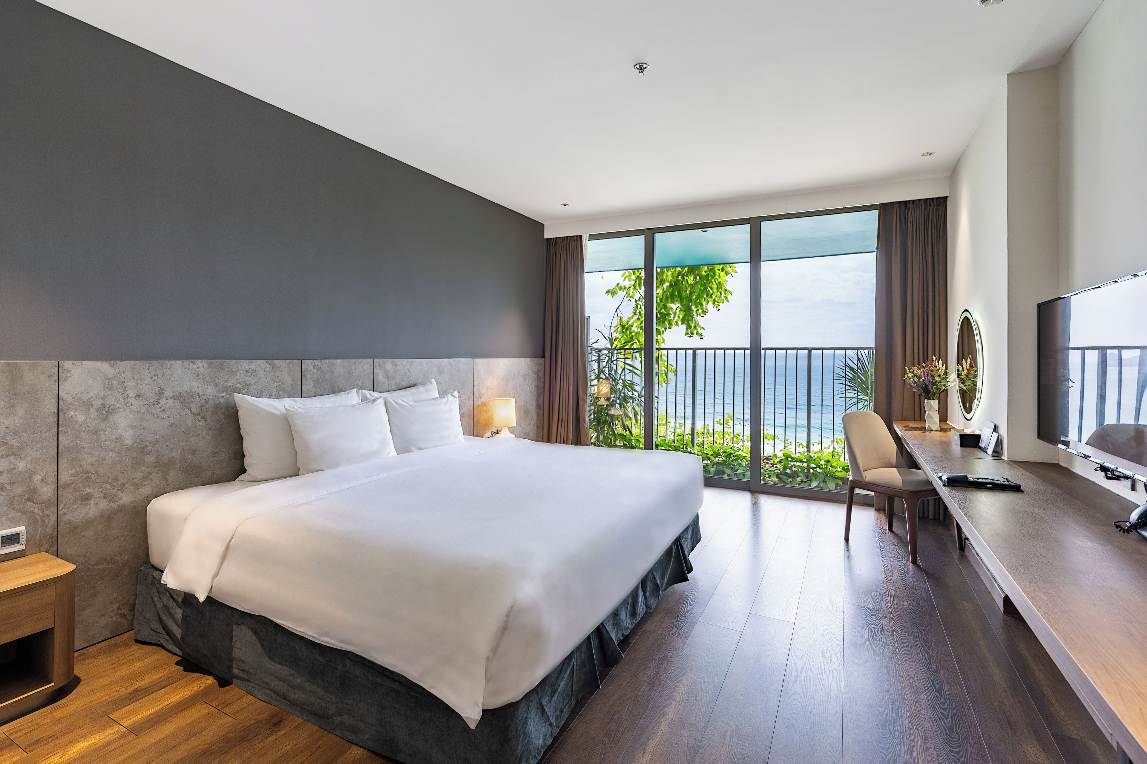 A modern hotel room with a large bed, a desk with a chair, a TV, and a balcony with a view of the sea.