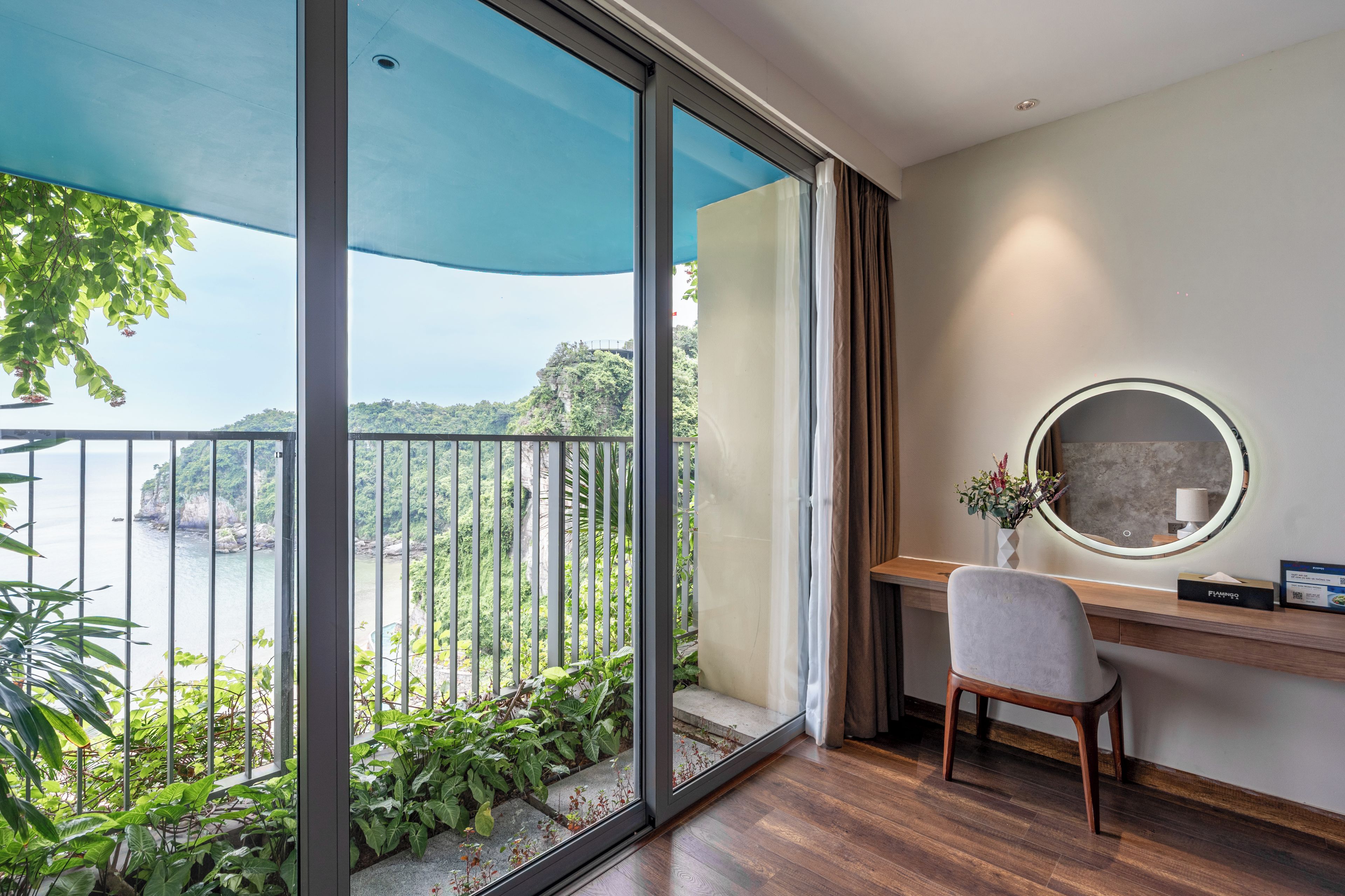 Une pièce comprenant un bureau, une chaise et une grande fenêtre donnant sur un balcon agrémenté de verdure et offrant une vue sur une falaise.