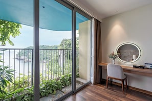 Una habitación con escritorio, silla y un ventanal grande que da a un balcón con vegetación y vistas a un acantilado.