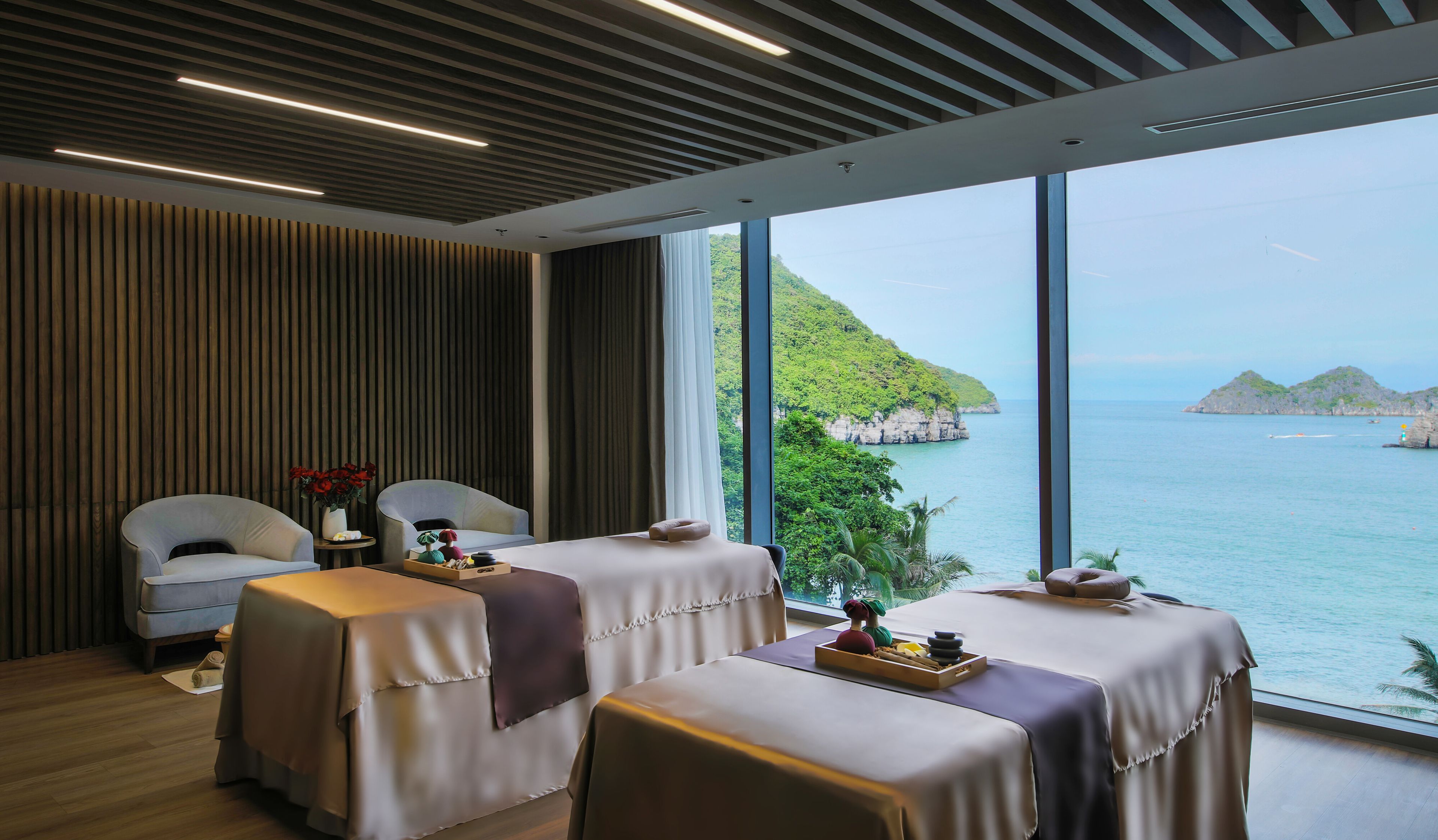 A spa room with two massage tables, a small table with a vase of flowers, and a view of the sea and hills.