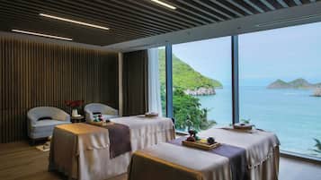 A spa room with two massage tables, a small table with a vase of flowers, and a view of the sea and hills.