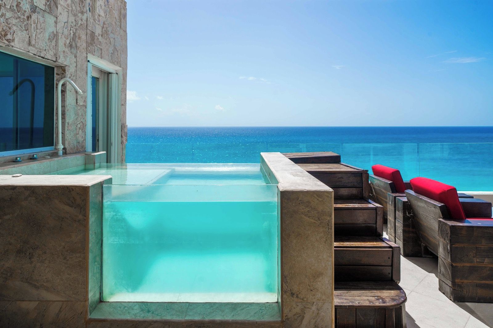 A rooftop infinity pool with clear blue water, wooden deck seating, and a view of the ocean.