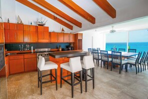 Cozinha moderna com armários de madeira, uma ilha central com banquetas e uma área de refeições com vista para o oceano.