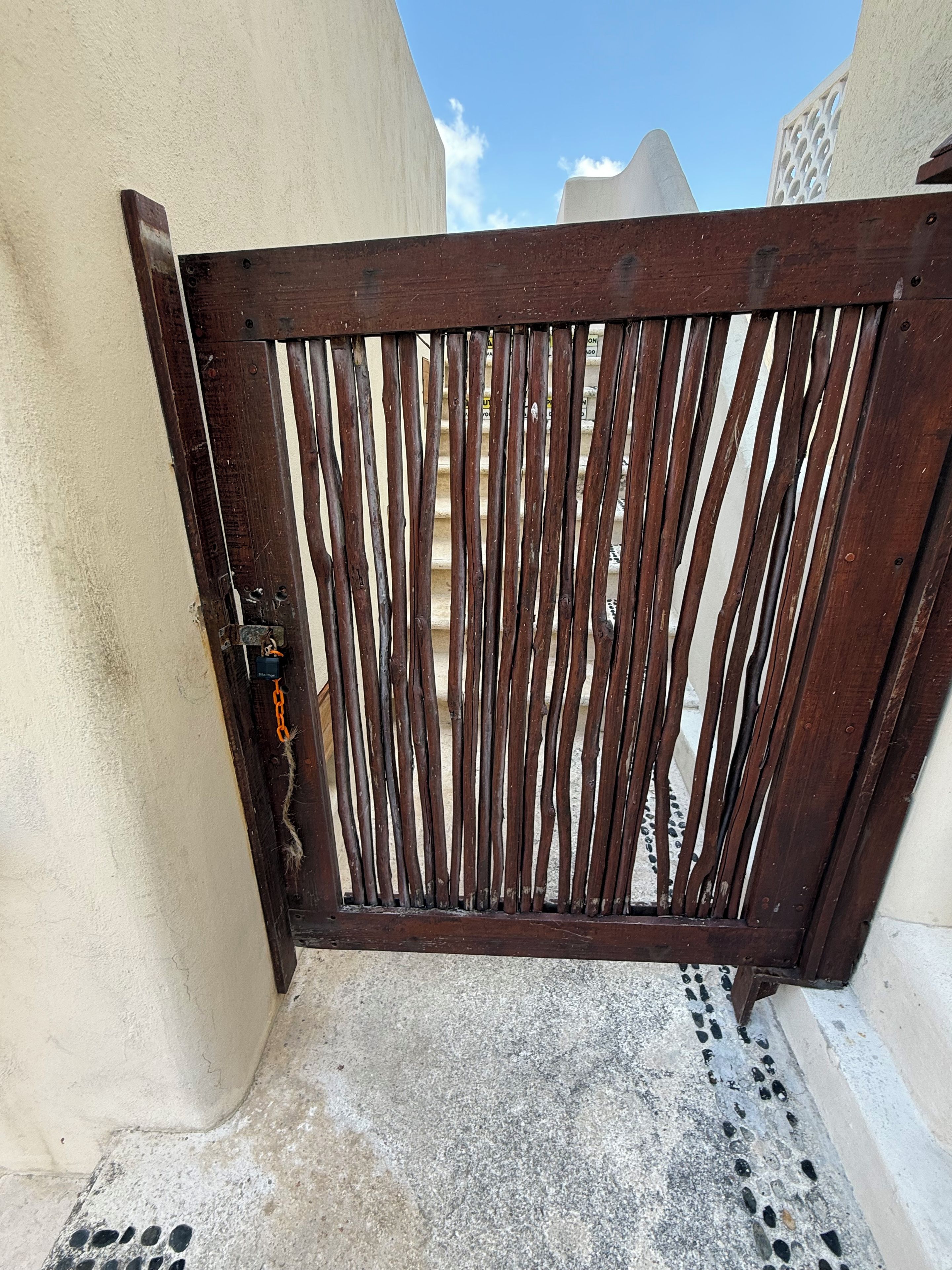 Una porta de fusta amb llistons verticals, un cadenat i un camí de formigó que porta a un edifici amb façana blanca.