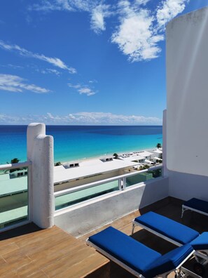 A balcony with a view of the ocean and beach, wooden flooring, and blue lounge chairs. - Luxury Oceanfront Suite with Jacuzzi (Cancún)
