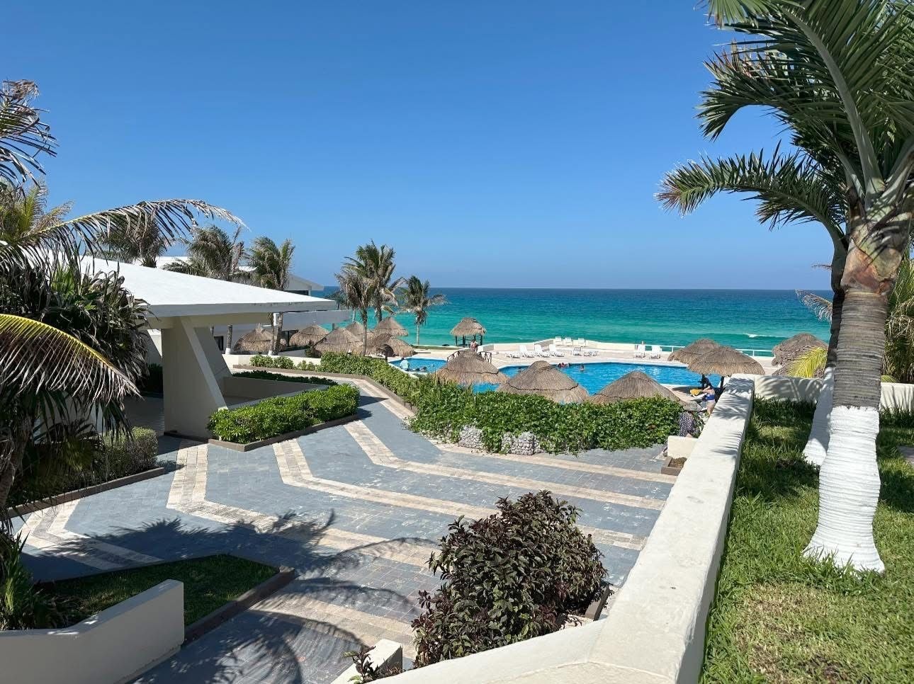 Zona de piscina en un resort con vista despejada al océano, rodeada de palmeras y sombrillas de paja.