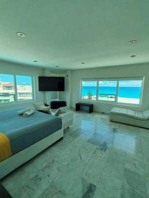 A modern bedroom with a bed, a TV, a seating area, and large windows with a view of the sea. - Luxury beachfront suite with a hot tub (Cancún)