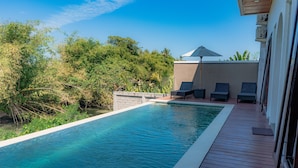 Uma piscina retangular com um deck de madeira ao redor, algumas cadeiras de descanso e um edifício com fachada branca e telhado inclinado.