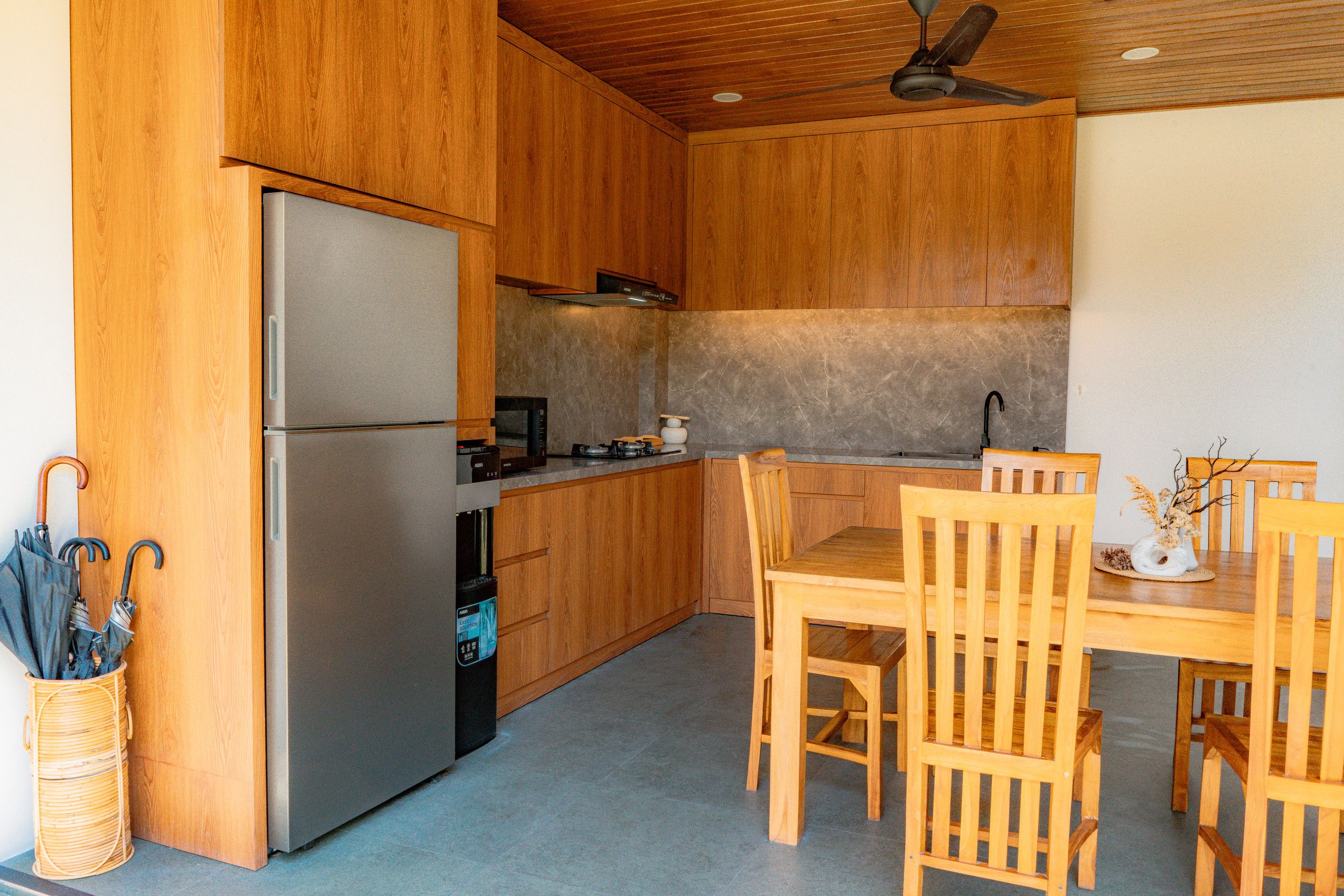 Cozinha compacta com armários de madeira, geladeira de aço inoxidável e área de refeições com cadeiras e mesa de madeira.