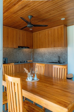 Een houten interieur met een eettafel, stoelen, een plafondventilator en een keuken met aanrecht en spoelbak.