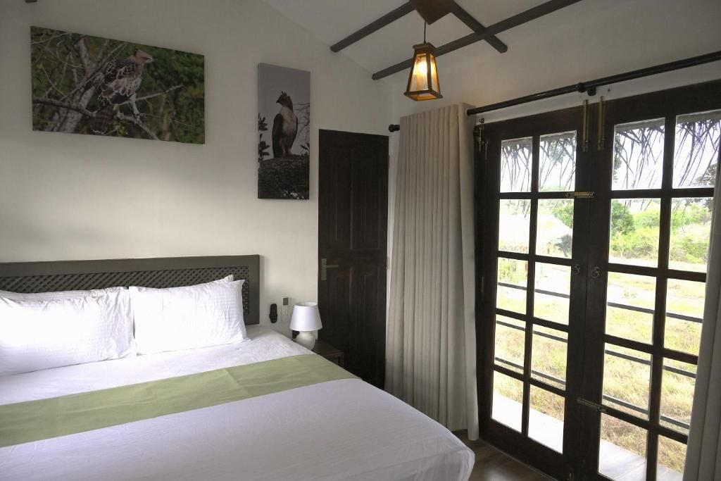 Una habitación de hotel con cama, mesita de noche, lámpara, ventana con cortinas y puerta.