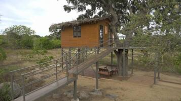 A wooden cabin with a thatched roof, a wooden staircase leading up to it, and a bench outside.