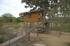 Eine Holzhütte mit einem Strohdach, eine Holztreppe führt hinauf zu ihr und eine Bank steht davor.