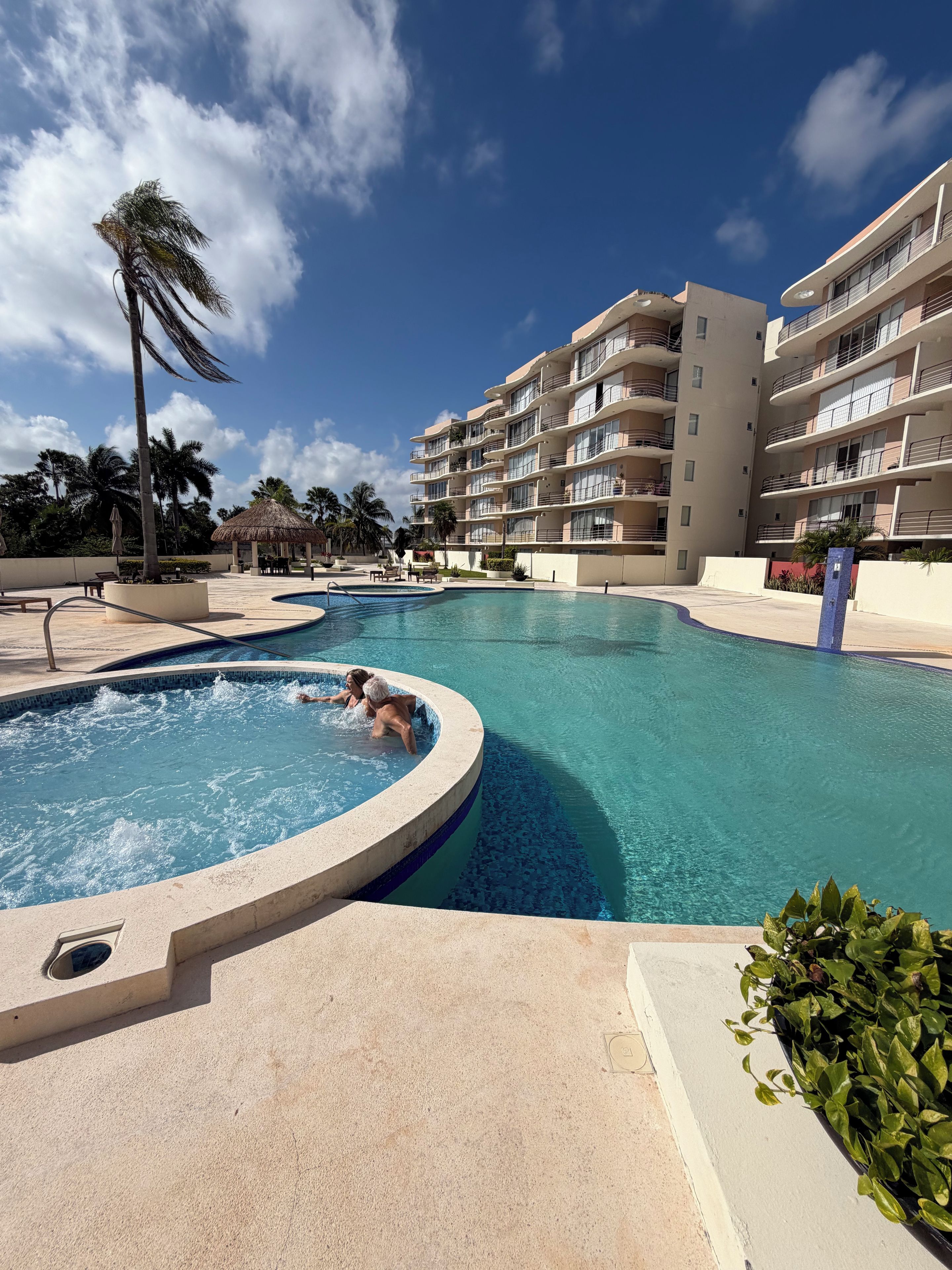 A swimming pool area with a hot tub, palm trees, and a multi-story building in the background.
