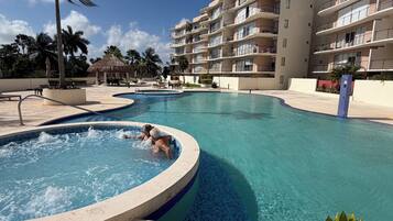 A swimming pool area with a hot tub, palm trees, and a multi-story building in the background.