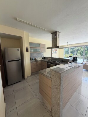 A modern kitchen with a stainless steel refrigerator, granite countertops, wooden cabinets, and a large window providing a view of greenery outside. - Luxury apartment with a balcony in the Cumbres shopping plaza (Cancún)