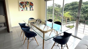 Een moderne woonkamer met een glazen eettafel, zwarte stoelen, een bank en een balkon met uitzicht op groen.
