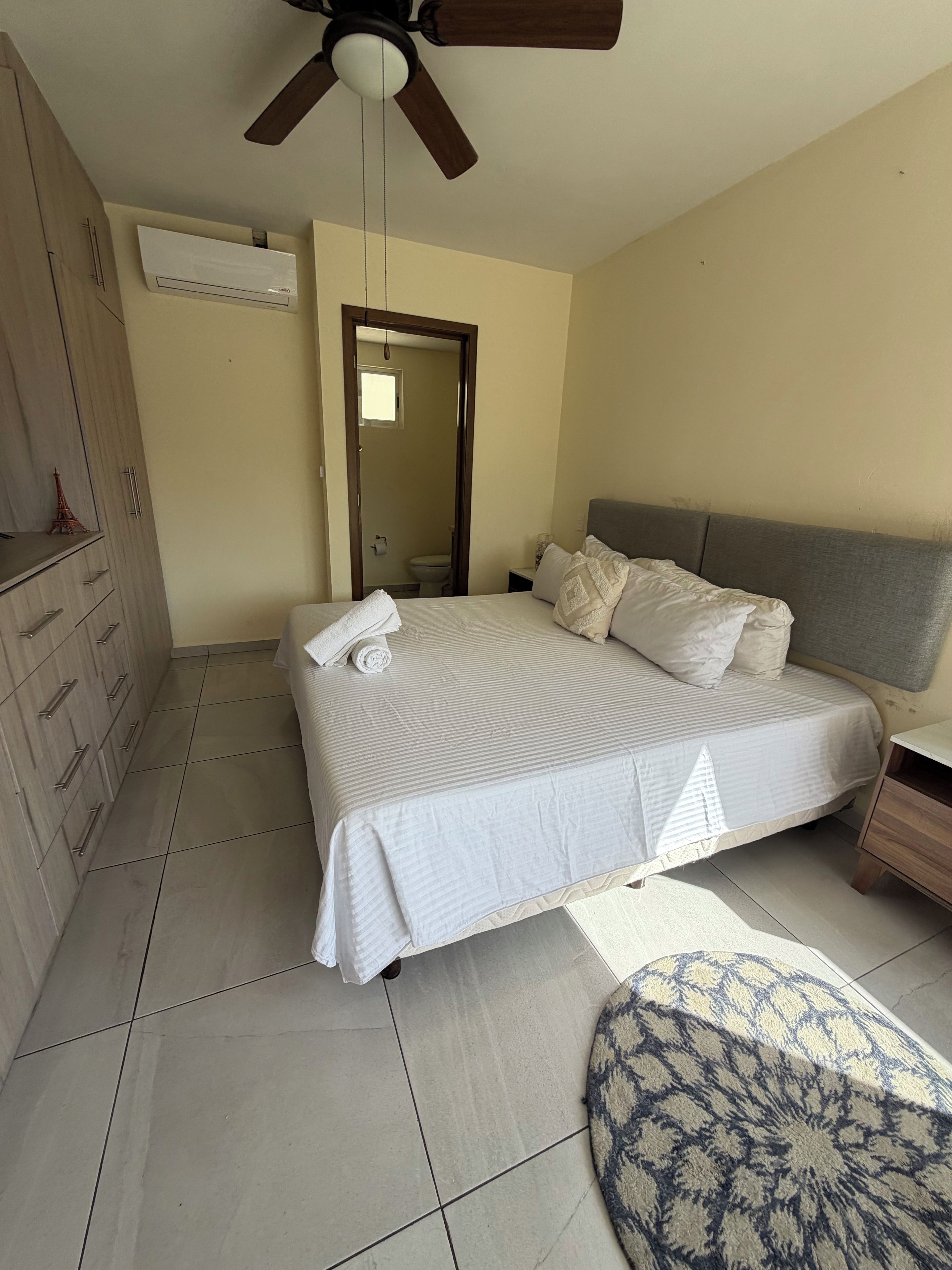 A bedroom with a bed, a wardrobe, a ceiling fan, and a bathroom visible through an open door.