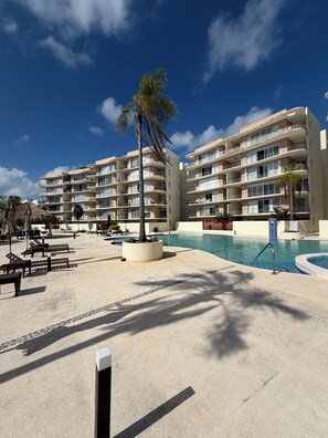 A resort with a swimming pool, lounge chairs, and a palm tree. - Luxury apartment with a balcony in the Cumbres shopping plaza (Cancún)