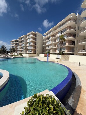 A multi-story hotel with a swimming pool, balconies, and palm trees. - Luxury apartment with a balcony in the Cumbres shopping plaza (Cancún)