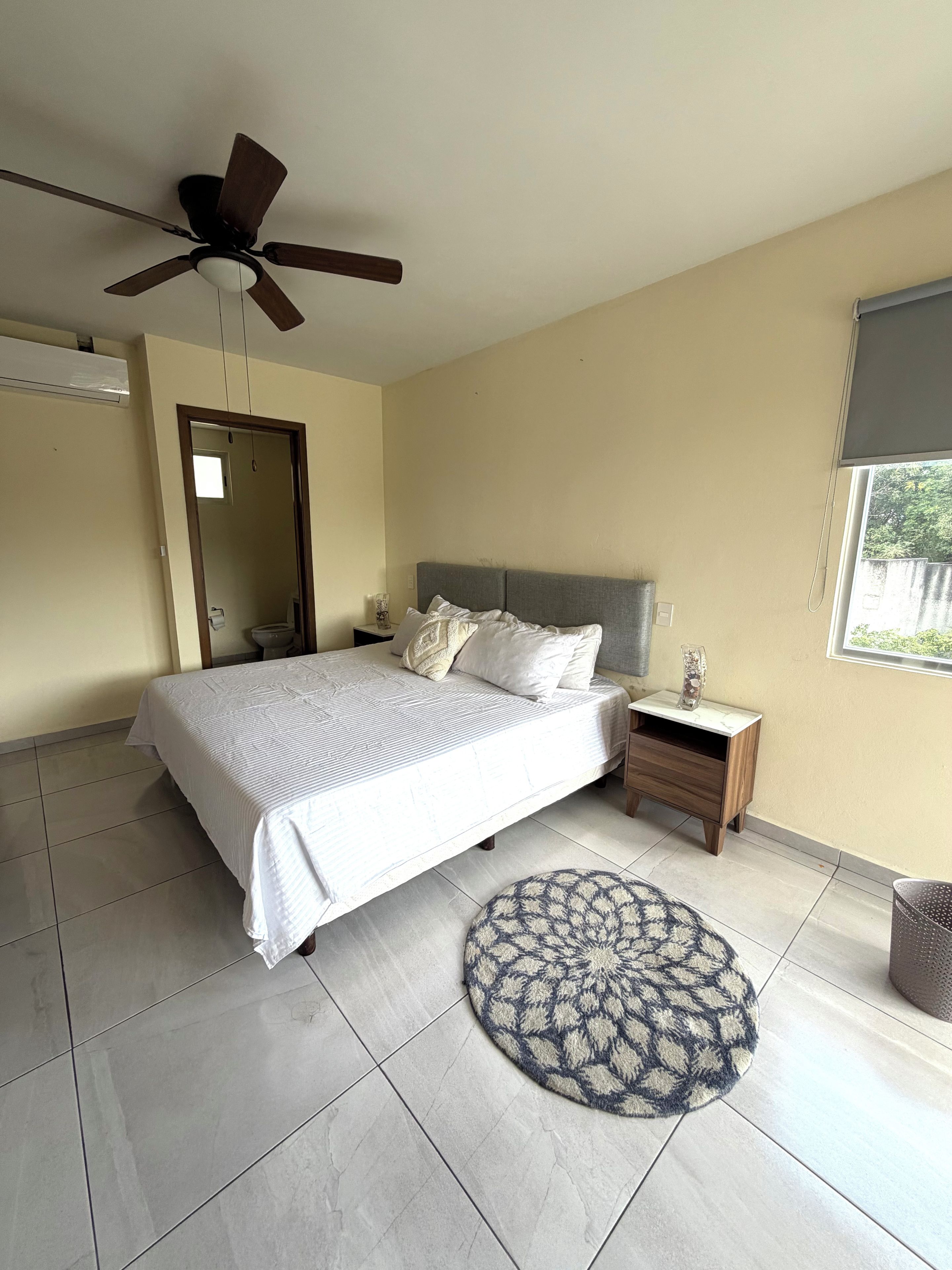 A bedroom with a bed, a nightstand, a bedside table, a round rug, a ceiling fan, a window with a view, and an air conditioning unit.