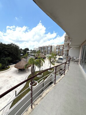 A balcony with a view of a resort area, including a pool, palm trees, and a thatched-roof structure. - Luxury apartment with a balcony in the Cumbres shopping plaza (Cancún)