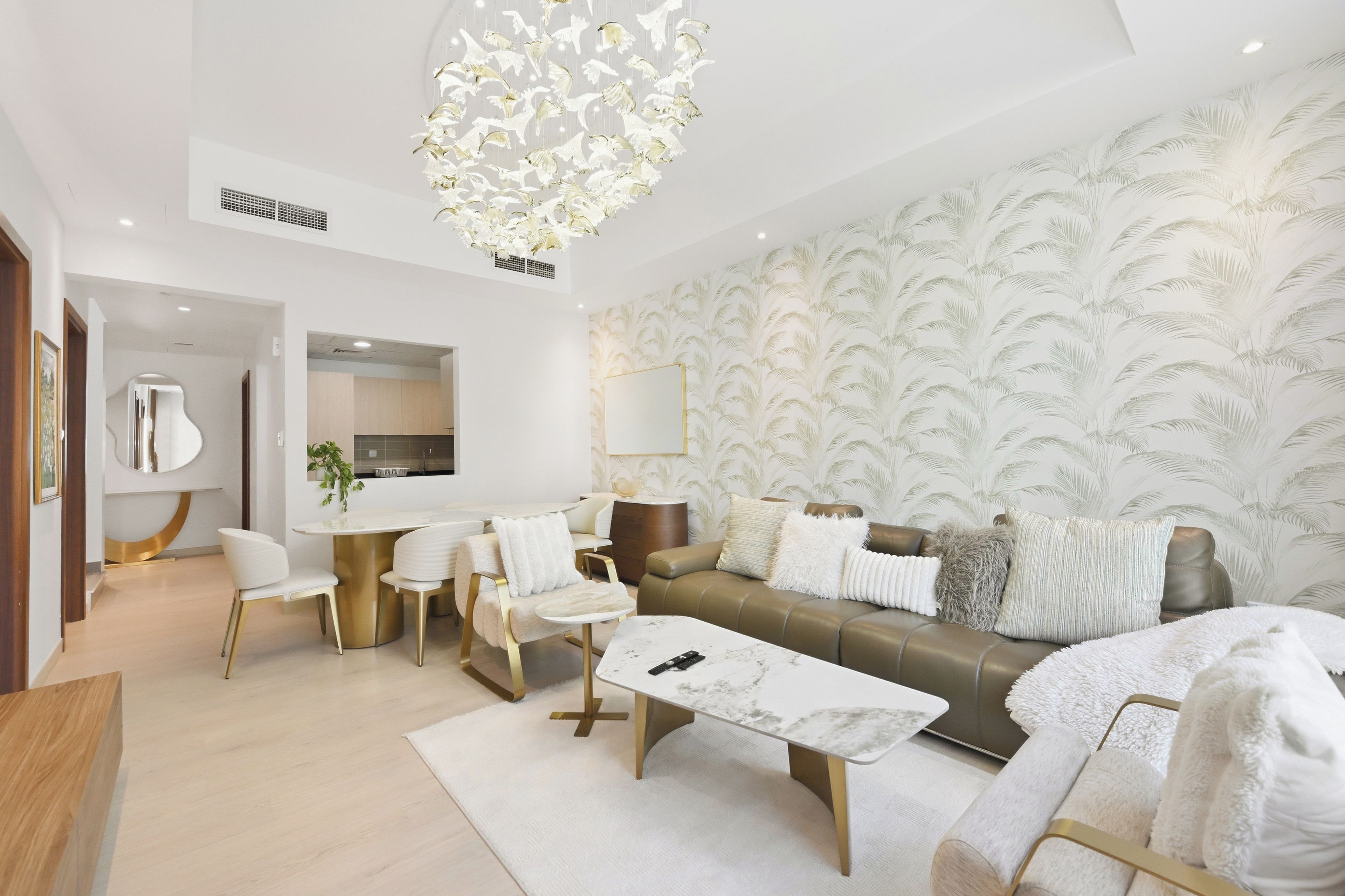 A modern living room with a sofa, coffee table, and chairs. There is a dining area with a table and chairs. The room has a large mirror and a decorative ceiling light.