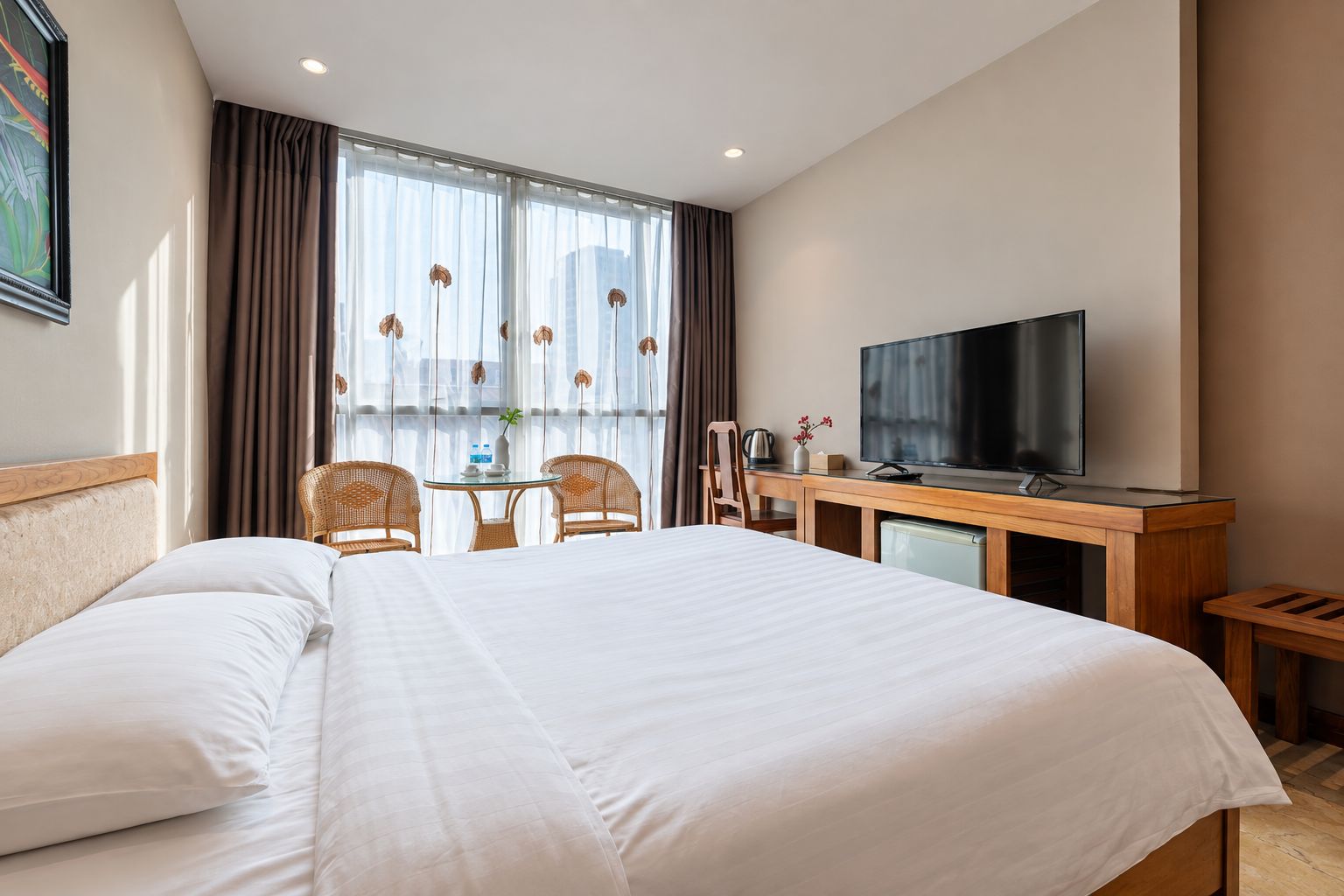 A hotel room with a large bed, a desk with a television, two wicker chairs, a small table, and a window with curtains.