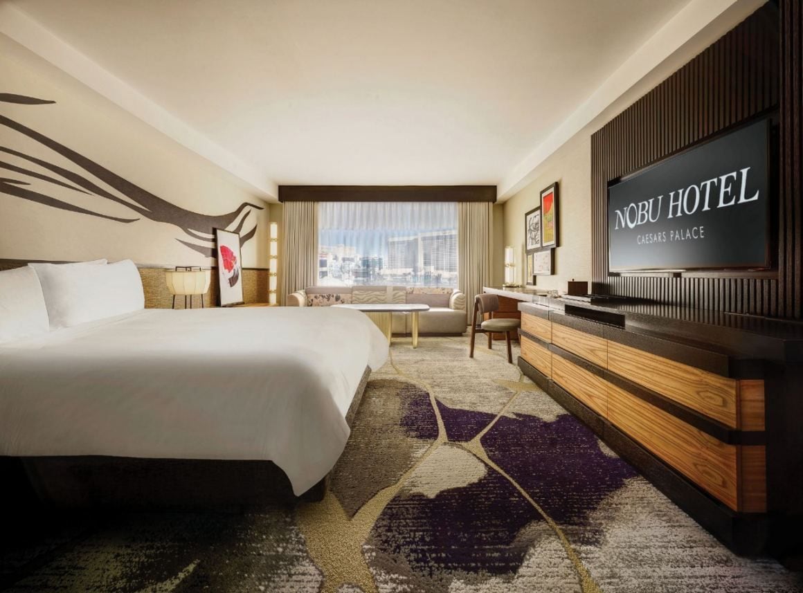 A hotel room with a bed, a desk, a television, and a view of the city.